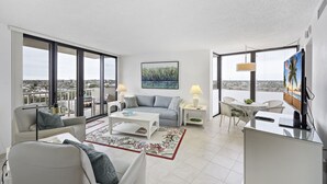 A modern living room with a large window, a sofa, a coffee table, and a TV. - Beachfront Condo | Pool, Tennis & Fitness Center (Marco Island)