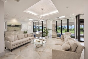 A spacious living room with a beige sofa, a glass coffee table, and a modern chandelier. - Beachfront Condo | Pool, Tennis & Fitness Center (Marco Island)