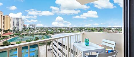 A balcony with a table and chairs, overlooking a tennis court and parking garage.