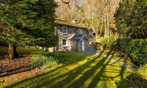 Une maison en pierre avec une cheminée en pierre, entourée d’une végétation luxuriante et d’arbres.