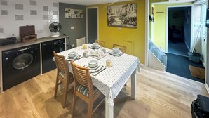 Un comedor con una mesa dispuesta para cuatro, sillas de madera, una lavadora y un ventilador al fondo.
