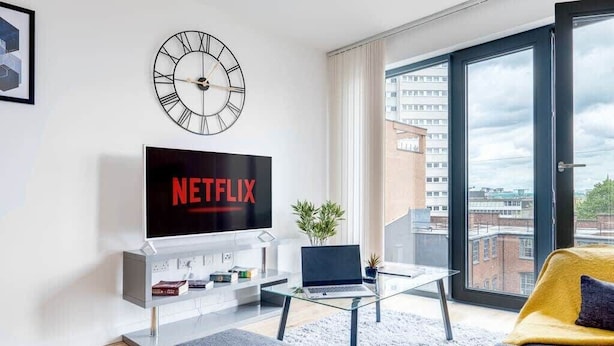 A modern living room with a large flat-screen TV displaying 'NETFLIX', a glass coffee table, a laptop, and a sofa with a yellow cushion. - Apollo Luxury - Netflix - Fast WIFI (West Midlands)