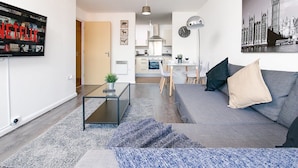 Un soggiorno moderno con un divano grigio, un tavolino da caffè e una TV che mostra contenuti Netflix. Si intravede una cucina sullo sfondo con un'area pranzo.