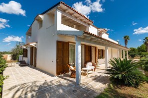 A two-story house with a covered patio, wooden doors, and a garden with palm trees.