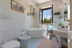 Casa de banho com banheira, duas casas de banho de apoio, um bidé e janela com vista para vegetação.