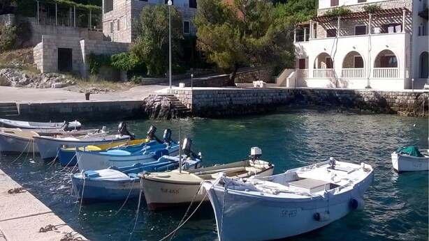 A marina with several boats docked, a stone building with a red roof, and a white building with arched windows. - Apartments Cumbelic (Mljet)