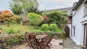 A wooden deck with a table and chairs, surrounded by a garden with various plants and a white building in the background. - Old Coach House (Ambleside)