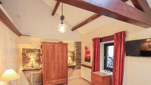 A bedroom with a bed, a wooden wardrobe, a stone wall, a radiator, a window with curtains, and a television on the wall. - Old Coach House (Ambleside)
