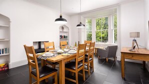 A dining area with a wooden table and chairs, a bookshelf, and a window with a view of greenery. - Ben House (Ambleside)