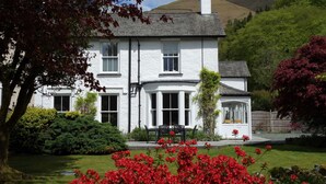 A white house with a bay window, surrounded by a well-maintained garden with red flowers and a lawn. - Ben House (Ambleside)