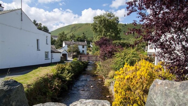 A narrow rural pathway flanked by white buildings and a stream, leading to a hill in the background. - Gable End (Keswick)
