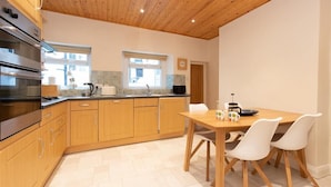 A modern kitchen with wooden cabinets, a stainless steel stove, and a dining area with a wooden table and white chairs. - Gable End (Keswick)