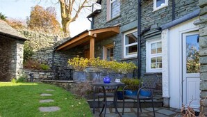 Uma casa de pedra com um pátio, incluindo uma mesa e cadeiras, cercada por um muro de pedra e um gramado.