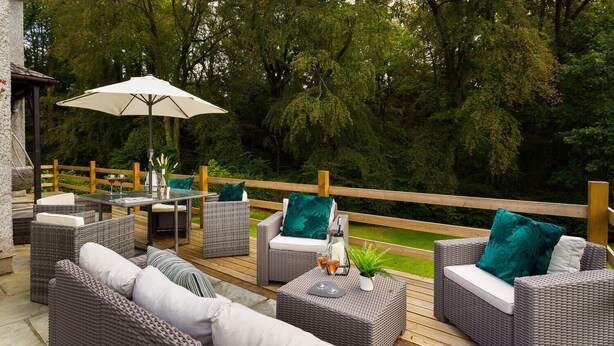 An outdoor patio with wicker furniture, a table with glasses, and a wooden deck overlooking a garden. - Beech View (Ambleside)