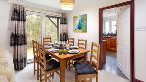A dining area with a wooden table set for six, surrounded by chairs with black cushions. A peacock painting hangs on the wall, and a window with curtains is visible. - Beech View (Ambleside)