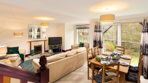 A modern living room with a sofa, TV, fireplace, dining table with chairs, and large windows overlooking a garden. - Beech View (Ambleside)