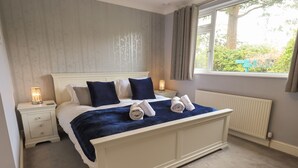 A neatly made bed with a blue blanket and two pillows, a bedside lamp, and a window with curtains. - Silver Birches (Windermere)