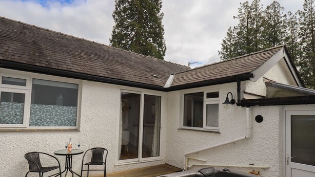 A hot tub on a wooden deck with two chairs and a table, a white building with windows, and a roof. - Silver Birches (Windermere)