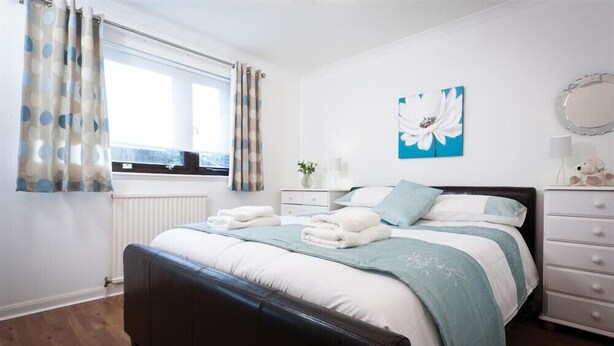 A bedroom with a bed, a nightstand, a dresser, a window with curtains, and a wall with a flower painting. - Mount Sol (Ambleside)