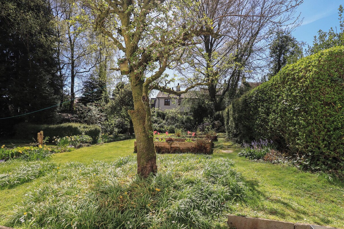 Un jardín con un árbol, parterres de flores y una casa al fondo.