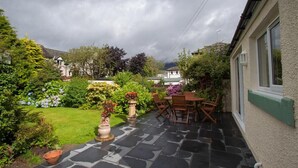 Un patio con suelo de piedra, mobiliario exterior y un jardín con diversas plantas y flores.