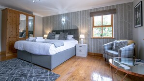 A bedroom with a large bed, bedside tables, a wardrobe, a window with a view, and a seating area with a chair and table. - Waltons Close (Penrith)