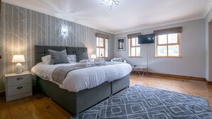 A bedroom with a large bed, bedside tables, a TV, and a window with curtains. - Waltons Close (Penrith)