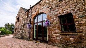 A stone building with a large arched doorway and a window with purple flowers. - Waltons Close (Penrith)