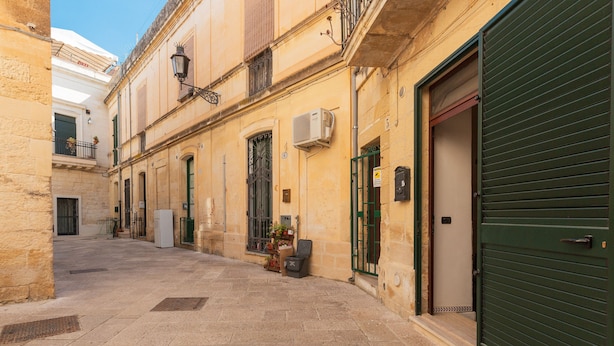 A narrow street with buildings on either side, featuring a closed green shutter door and a small shop with items displayed outside. - 2417 Dimora Santa Croce by Barbarhouse (Lecce)