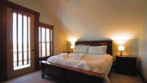 Een slaapkamer met een groot bed, twee nachtkastjes met lampen, een houten hoofdbord en een groot raam met jaloezieën.