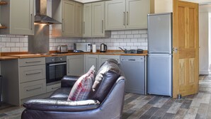Cocina compacta con gabinetes grises, extractor de acero inoxidable, horno integrado, lavavajillas y refrigerador. Un sofá de cuero con un cojín estampado se encuentra frente a la cocina.