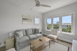 A living room with a grey sofa, a wooden coffee table, a white armchair, a ceiling fan, and a large window with a view of greenery and buildings.