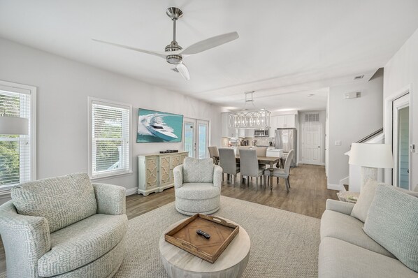 A modern living room with a ceiling fan, a dining area, and a kitchen in the background.