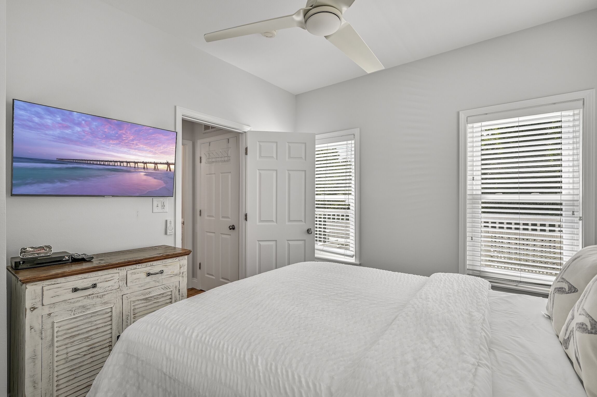 A bedroom with a bed, a ceiling fan, a TV on the wall, and a door leading to another room.