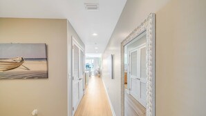 A hallway with wooden flooring, white walls, and doors with louvered shutters. A painting of a boat on a beach is visible on the left wall. - FREE Daily Golf! Reflections 211 Bayfront Beauty, Heated Resort Pool, Hot Tub (Panama City)