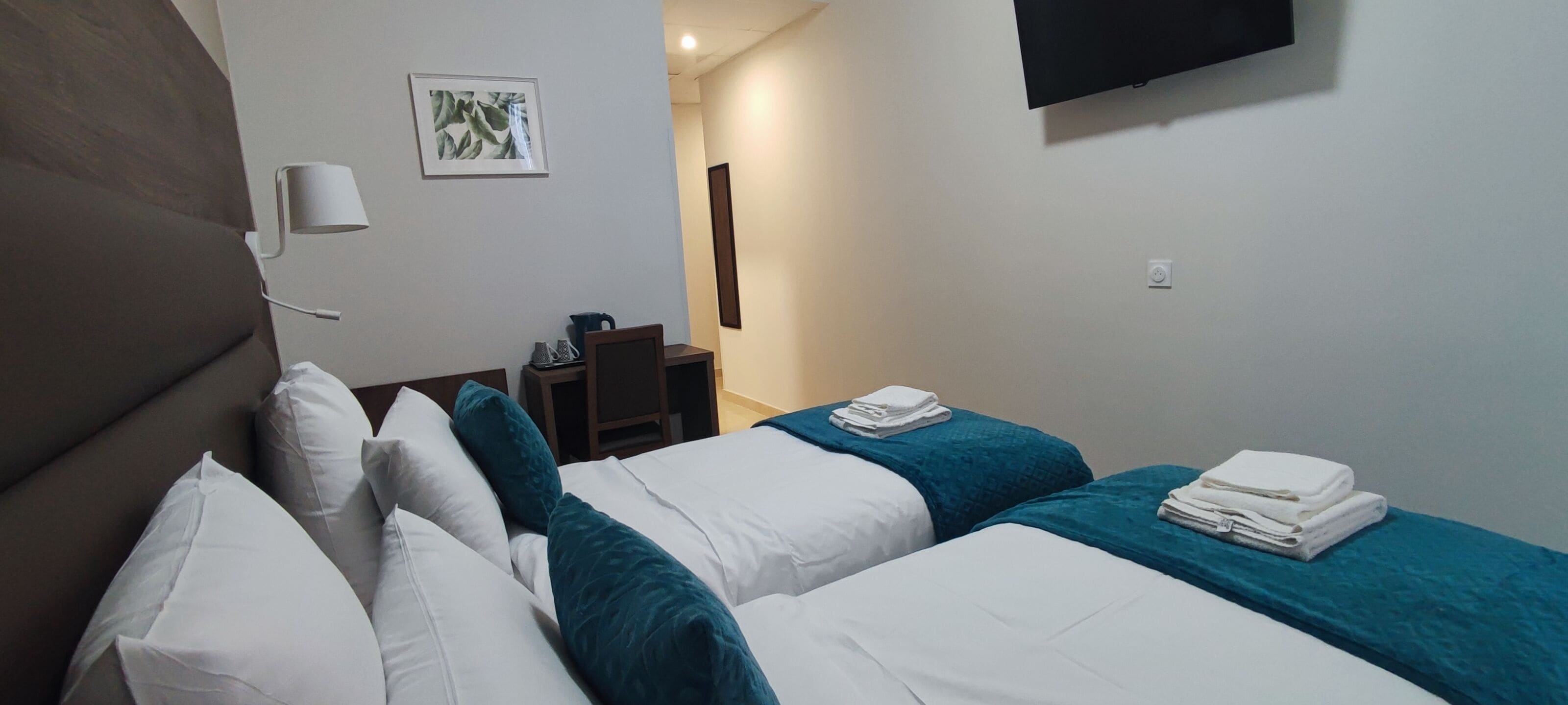 Een hotelkamer met twee bedden, een aan de muur bevestigde televisie, een nachtkastje met een lamp en een lijst aan de muur.