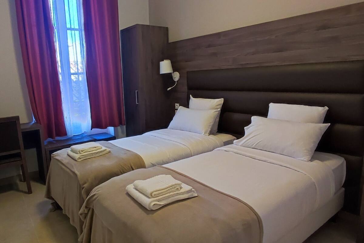 A hotel room with two beds, a desk, a wardrobe, and a window with curtains.