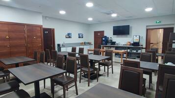 A hotel dining area with multiple tables and chairs, a coffee machine, and a television mounted on the wall.