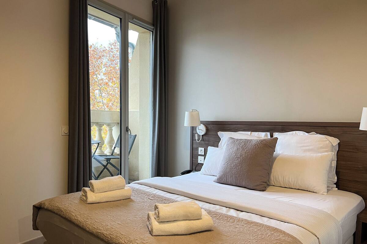 A hotel room with a large bed, two pillows, a bedside table with a lamp, and a view of a balcony with chairs.