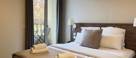Habitación de hotel con una cama grande, dos almohadas, una mesita de noche con lámpara y vistas a un balcón con sillas.