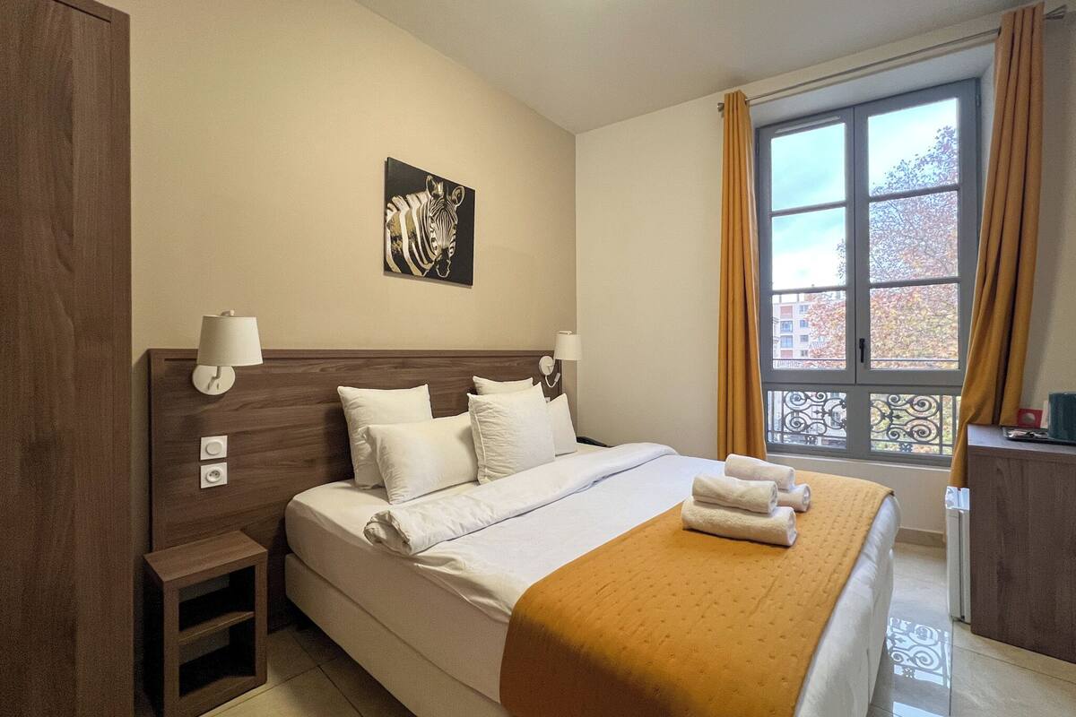 A hotel room with a large bed, a nightstand, a wall-mounted lamp, a window with curtains, and a zebra artwork on the wall.