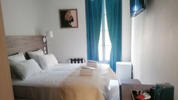 Una cama bien hecha con ropa de cama blanca, una mesita de noche con una lámpara, un cuadro en la pared y una ventana con cortinas azules.