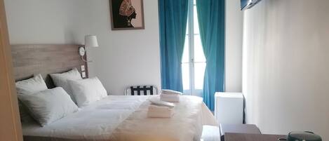 Een netjes opgemaakt bed met wit beddengoed, een nachtkastje met een lamp, een ingelijst schilderij aan de muur en een raam met blauwe gordijnen.