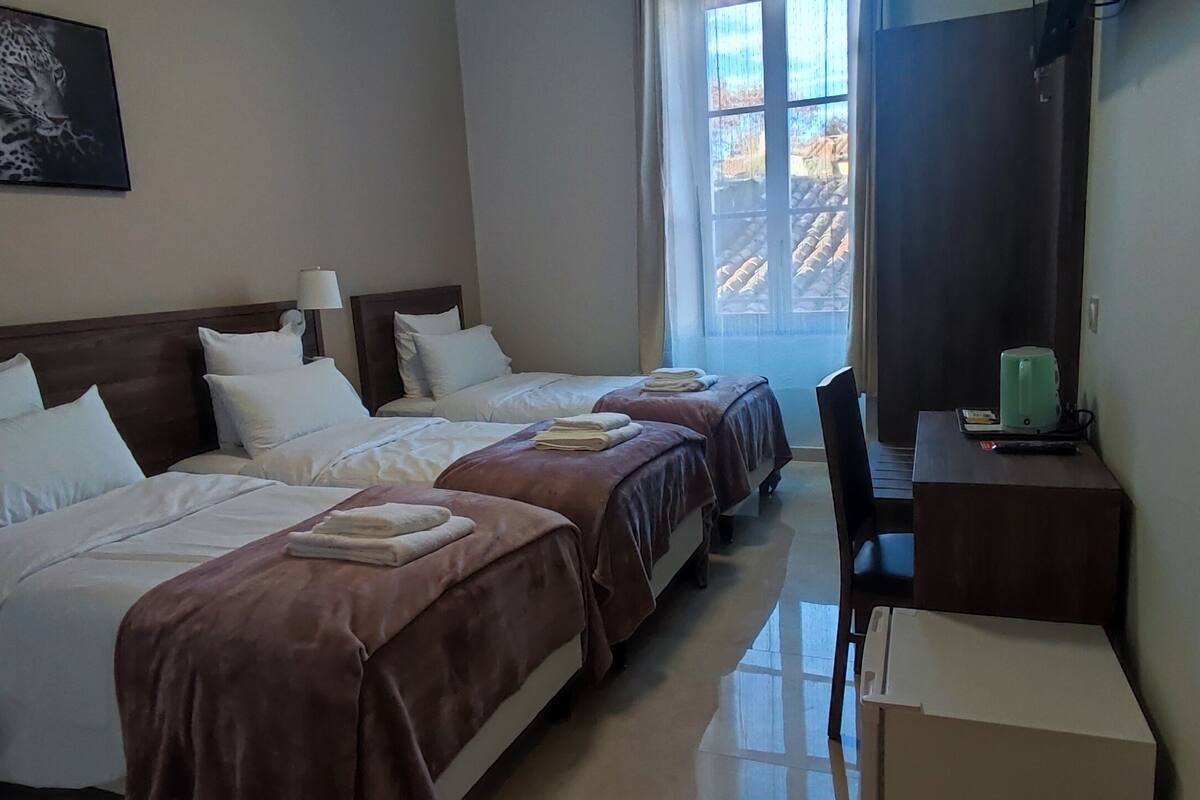 A hotel room with three beds, a desk with a kettle, and a window with a view of the outside.