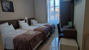 A hotel room with three beds, a desk with a kettle, and a window with a view of the outside.