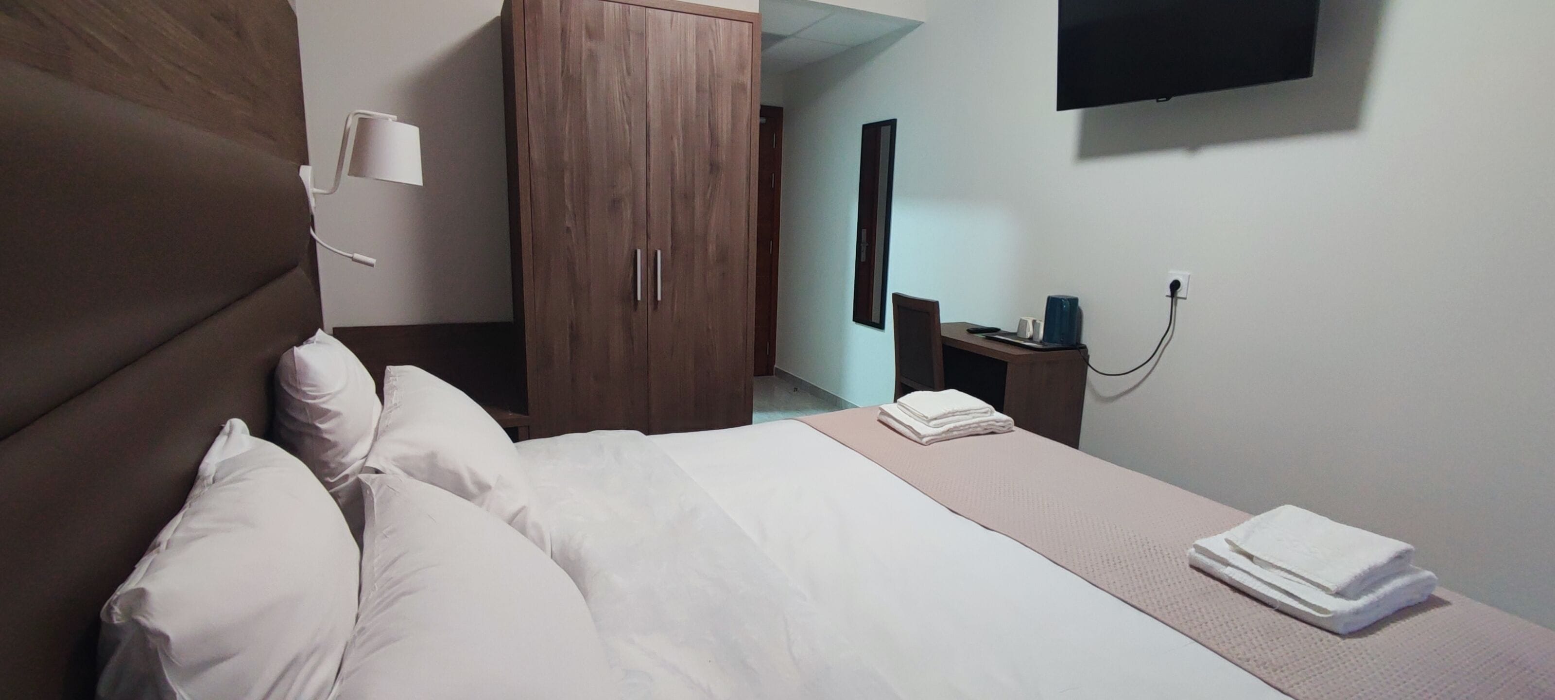 Habitación de hotel con cama, armario, escritorio con silla, televisor montado en la pared y mesita de noche con lámpara y teléfono.