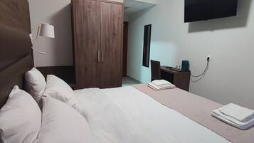 Habitación de hotel con cama, armario, escritorio con silla, televisor montado en la pared y mesita de noche con lámpara y teléfono.