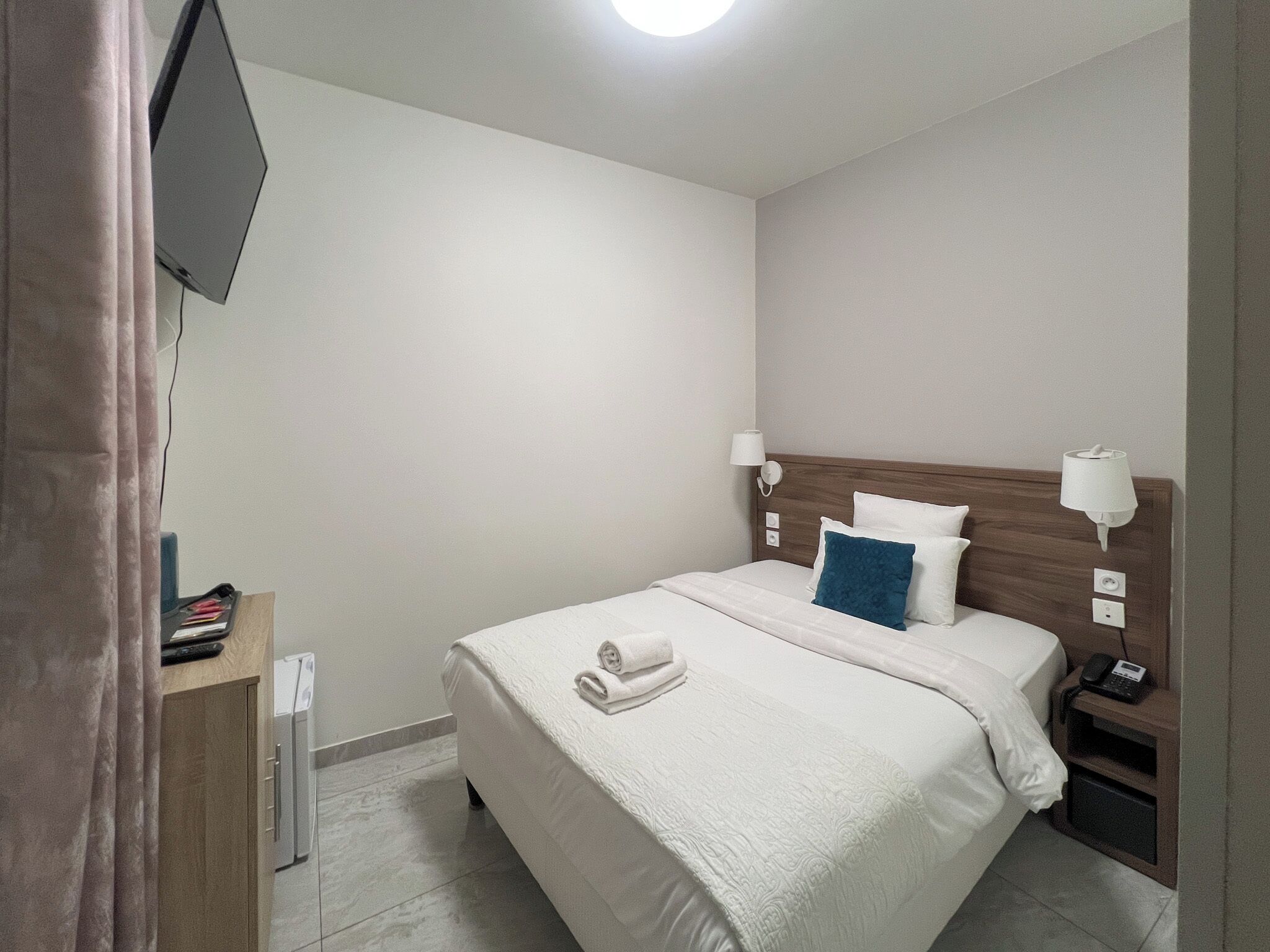 Habitación de hotel ordenada con una cama, lámparas de noche, teléfono en la mesita y un televisor montado en la pared.