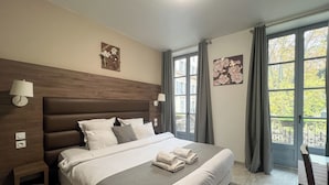 A hotel room with a large bed, a wooden headboard, two wall-mounted lamps, and a view of trees through the window. - Square Hôtel (Nîmes)