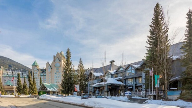 A snow-covered street with buildings, including a hotel with a green roof and a large building with a tower. - MAGNIFICENT SKI-IN/OUT 1BR: AC and Gym (Whistler)